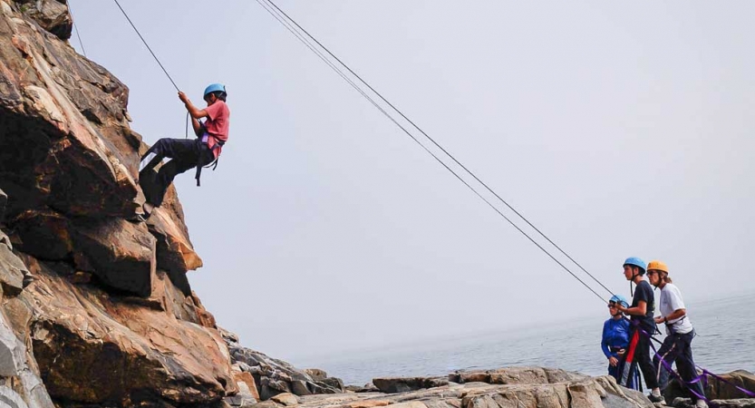 One person wearing safety gear is secured by ropes as they rappel a rock face. Others on the ground belay the climber. 